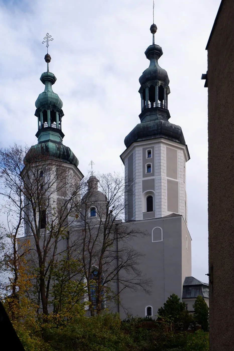 Dwie wieże kościoła z zielonymi, miedzianymi hełmami o barokowych, cebulastych kształtach, widziane z wąskiej uliczki pomiędzy drzewami i ciemnym murem