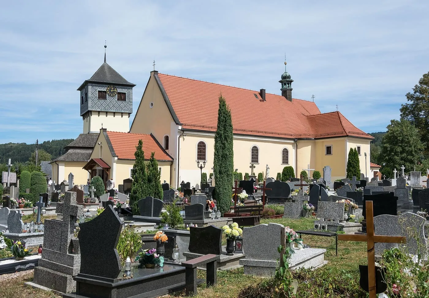 Jasnożółty kościół z czerwonym dachem i drewnianą dzwonnicą pod gontowym daszkiem stoi za cmentarzem w słoneczny dzień, na tle zalesionych wzgórz