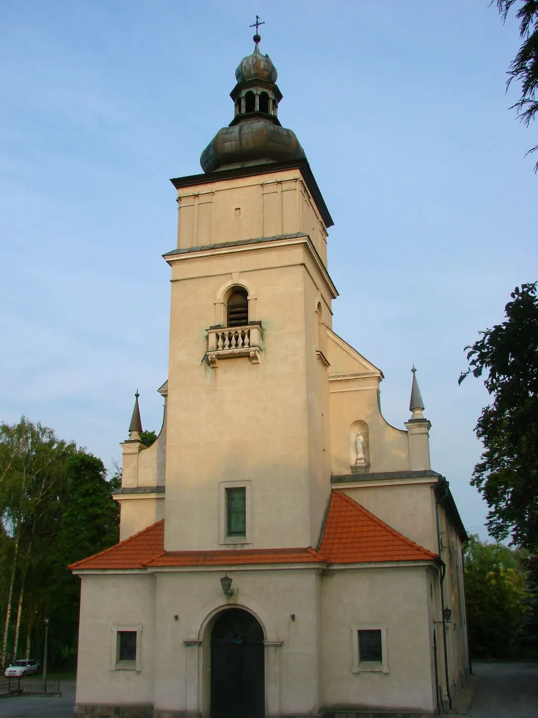 Jasny, klasycystyczny kościół z wysoką wieżą zwieńczoną baniastym hełmem i czerwonymi dachami nad przybudówkami, widziany od frontu