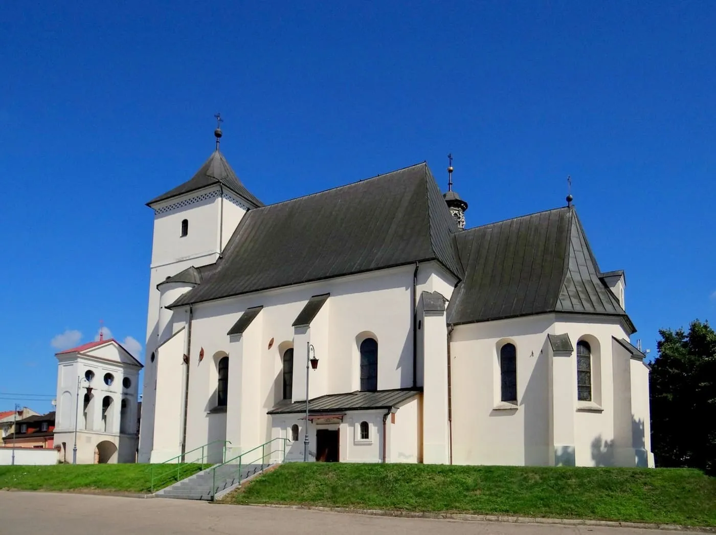 Biały, gotycki kościół św. Bartłomieja w Staszowie z przyporami, ciemnym dachem i czworoboczną wieżą, obok klasycystyczna dzwonnica, na tle błękitnego nieba