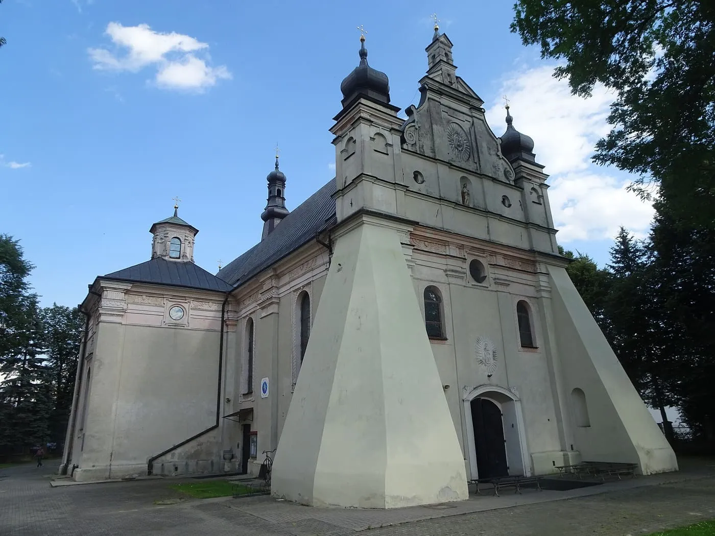 Biały, renesansowy kościół z wysokim szczytem fasady i dwiema smukłymi wieżyczkami zwieńczonymi ciemnymi hełmami, widziany z ukosa na tle błękitnego nieba z pojedynczą chmurą
