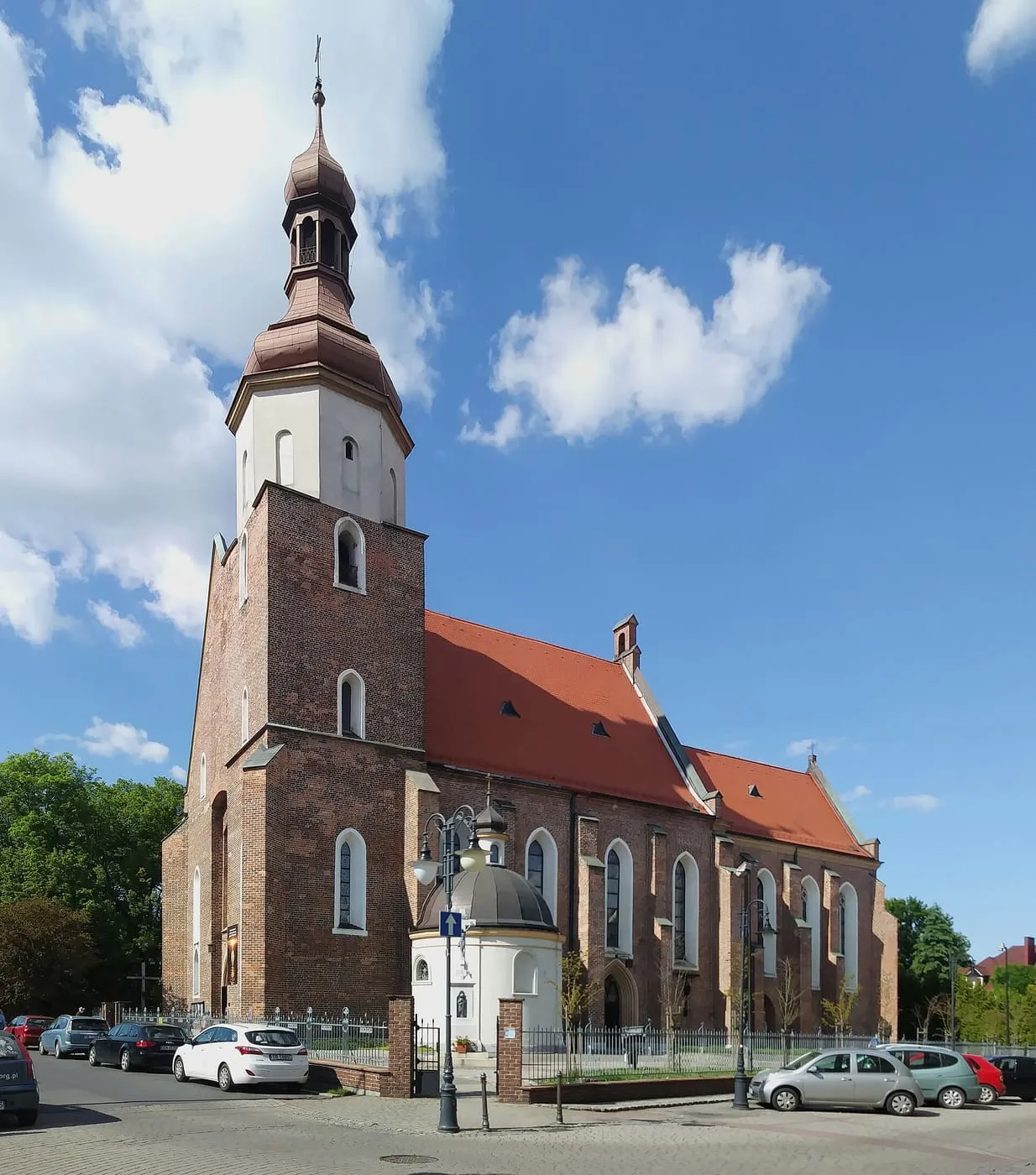 Ceglany, gotycki kościół farny w Żorach z wysoką wieżą zwieńczoną miedzianym, cebulastym hełmem oraz białą kaplicą dobudowaną u jej podstawy, widziany znad ulicy z zaparkowanymi samochodami