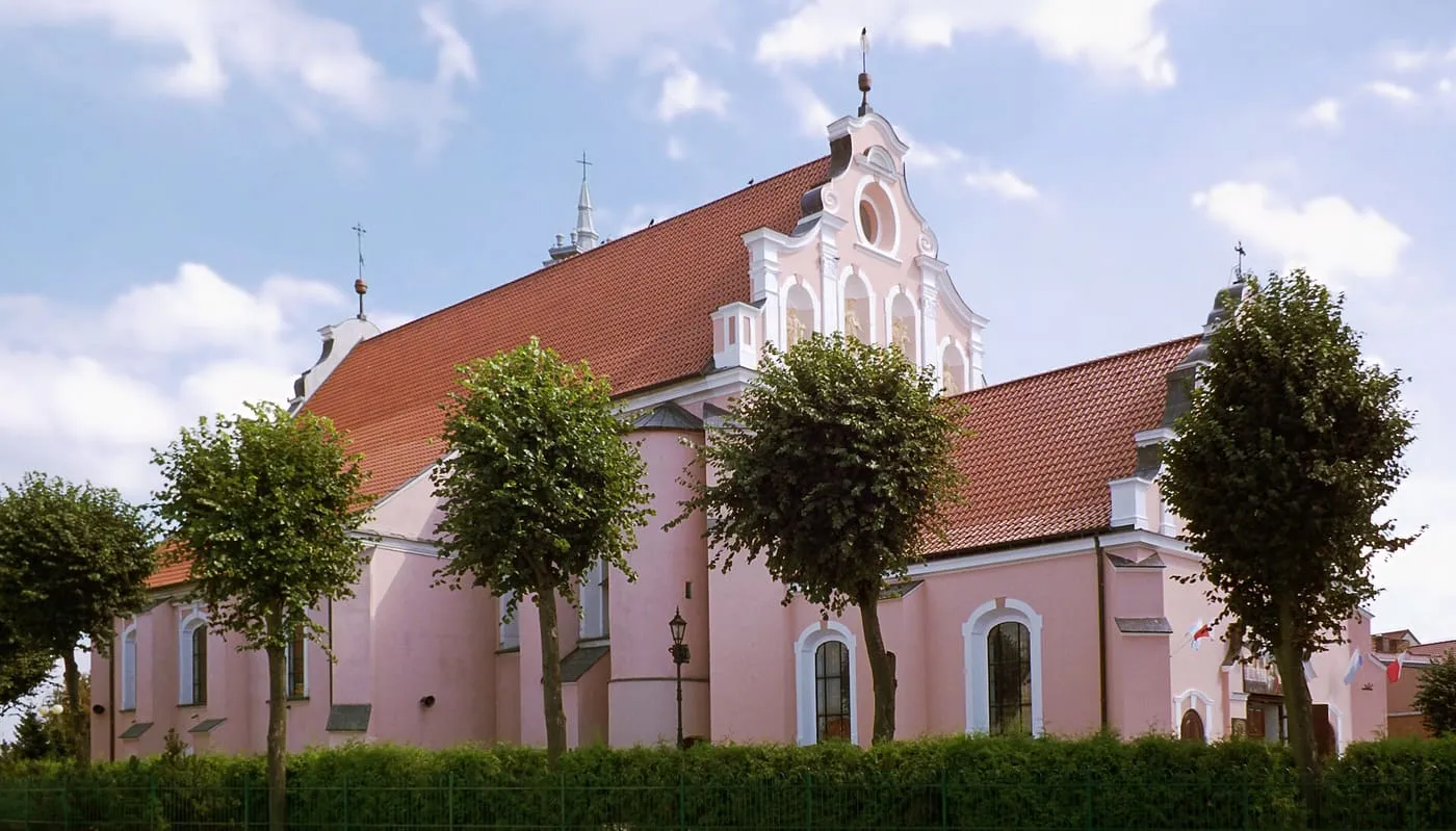 Różowy, barokowy kościół z czerwonym dachem i bogato zdobionym szczytem fasady, widziany z boku zza rzędu przystrzyżonych drzew i żywopłotu