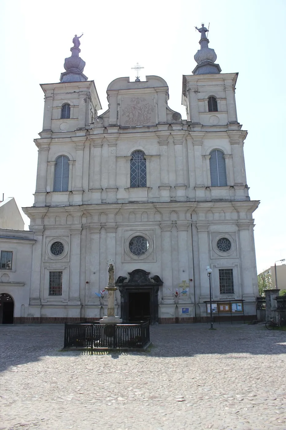 Symetryczna, biała barokowa fasada kościoła św. Franciszka Ksawerego w Krasnymstawie z dwiema wieżami zwieńczonymi figurami świętych i kapliczką z figurą Maryi na płycie rynku