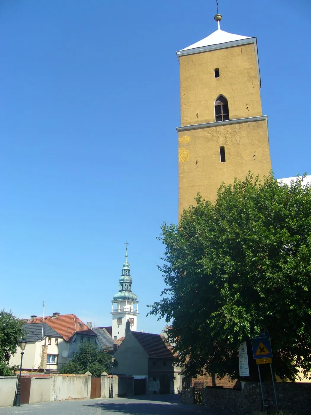 Czworoboczna wieża kościoła pokryta żółtawym tynkiem, z niskim białym dachem, a za nią wieża ratusza z zielonym hełmem, niżej domy i drzewo