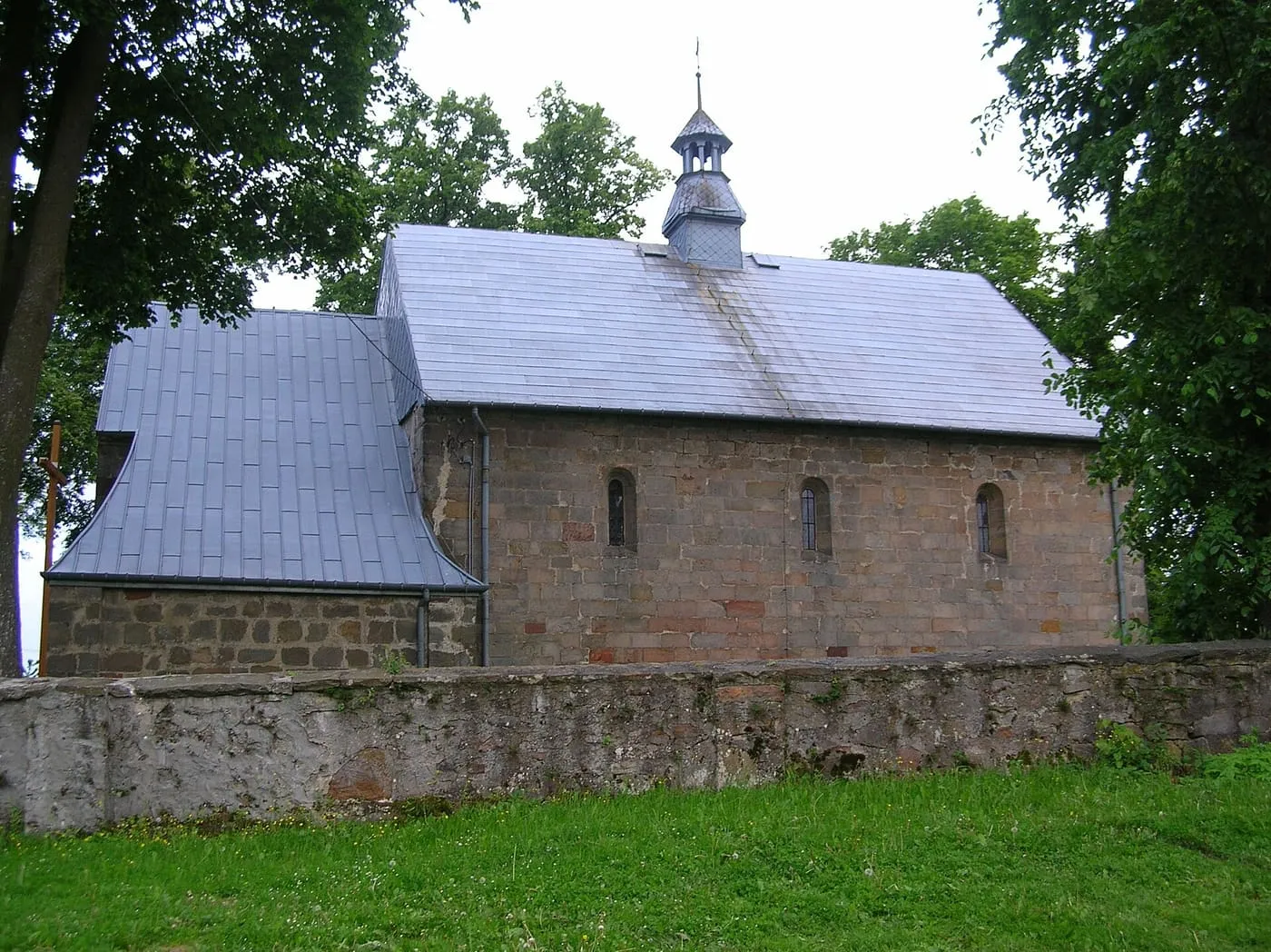 Mały, romański kościół z kamiennych ciosów w Tarczku widziany z boku, z wąskimi, półkoliście zamkniętymi oknami, szarym dachem i niewielką sygnaturką, za kamiennym murem wśród drzew