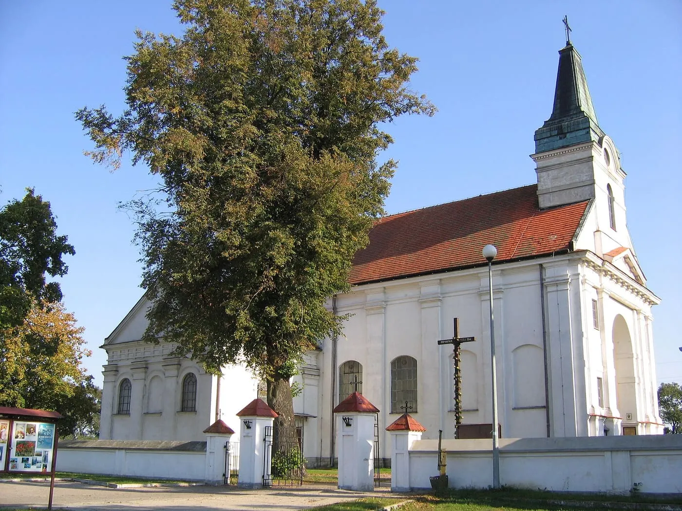 Biały, barokowy kościół z wieżą zwieńczoną zielonym hełmem, otoczony murem z bramkami i wysokim drzewem; na pierwszym planie krzyż z figurą Chrystusa