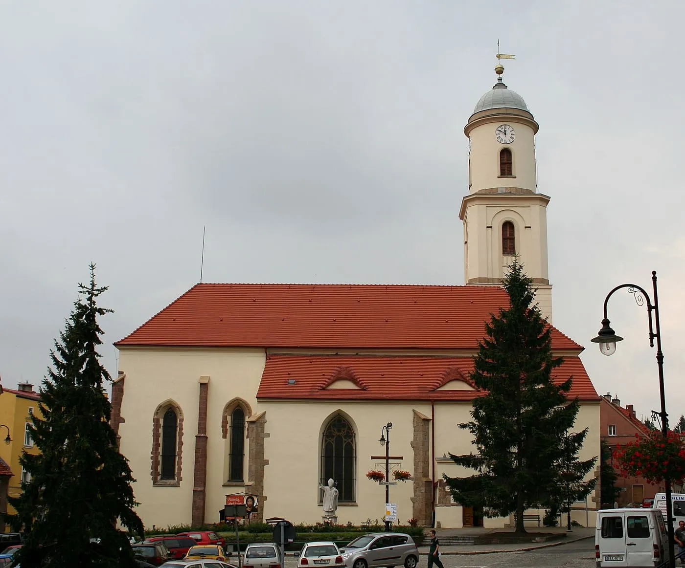 Kremowy kościół z czerwonym dachem i ostrołukowymi oknami, przy rynku; za nim biała wieża z zegarem i szarym hełmem, a na pierwszym planie choinki, latarnia i samochody