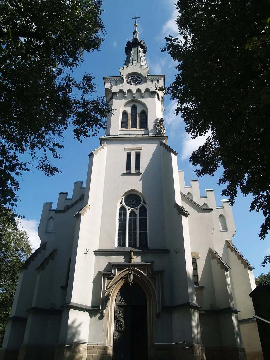 Biała fara św. Jadwigi w Dębicy widziana z dołu zza drzew, z wysoką wieżą zegarową zwieńczoną ciemną iglicą i ostrołukowym oknem z maswerkiem nad portalem