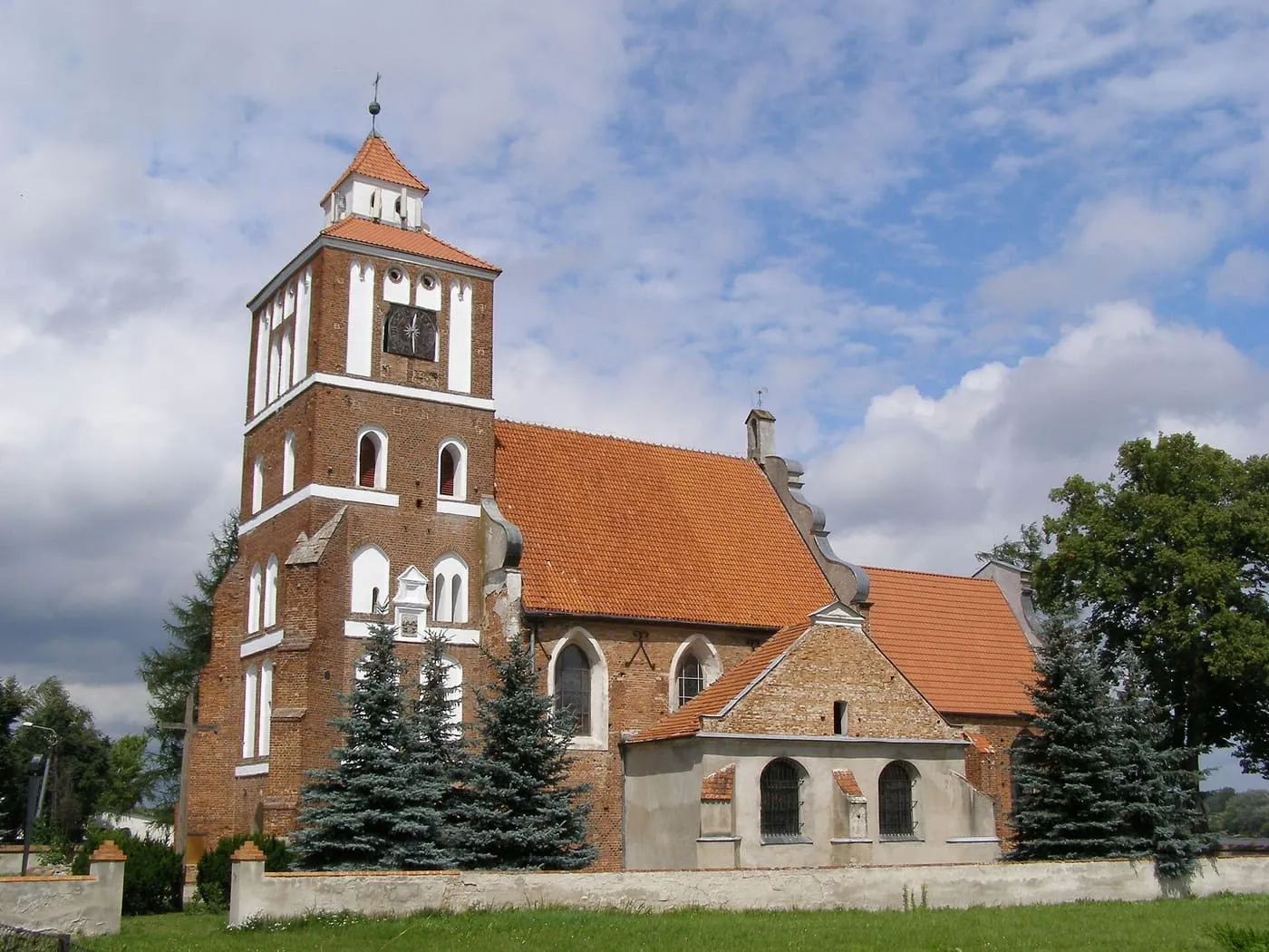 Ceglana wieża kościoła św. Jadwigi w Nieszawie z zieloną, namiotową iglicą, otoczona świerkami