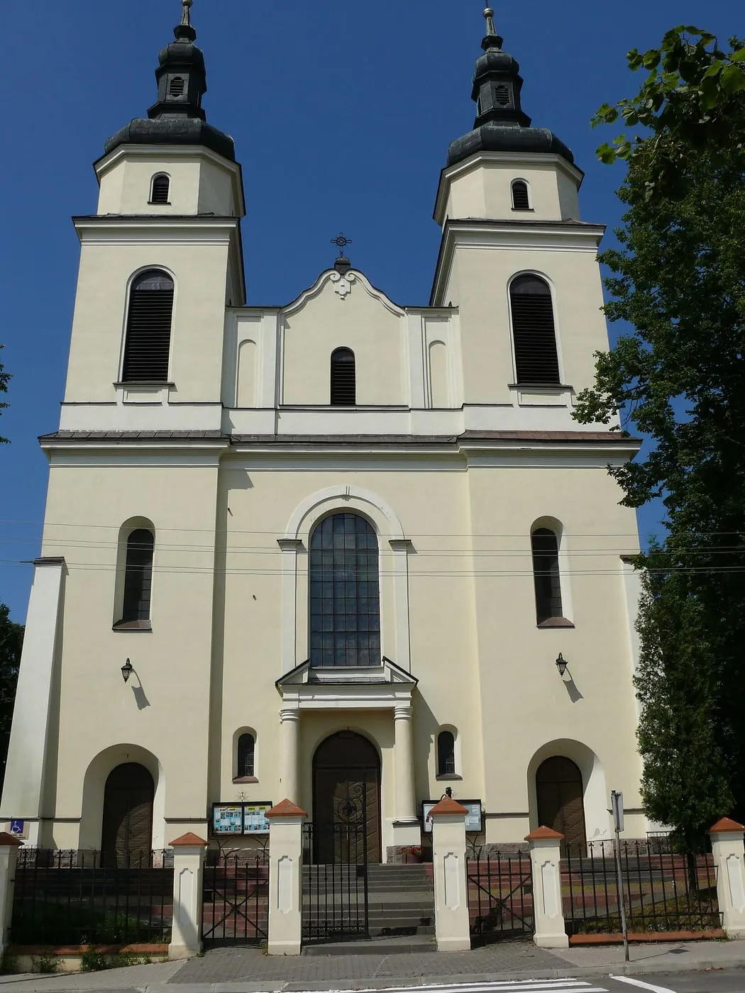 Jasny, neobarokowy kościół z dwiema symetrycznymi wieżami zwieńczonymi ciemnymi kopułami, za niskim murem i kutą bramą, na tle intensywnie błękitnego nieba
