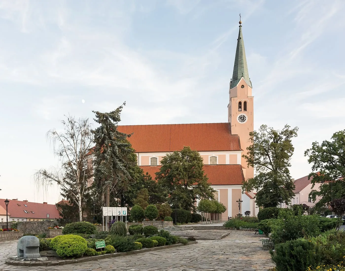 Kościół w kolorze łososiowym z czerwonym dachem i wysoką wieżą z zegarem i zielonym hełmem, częściowo zasłonięty drzewami nad ogrodzonym skwerem z kamienną nawierzchnią