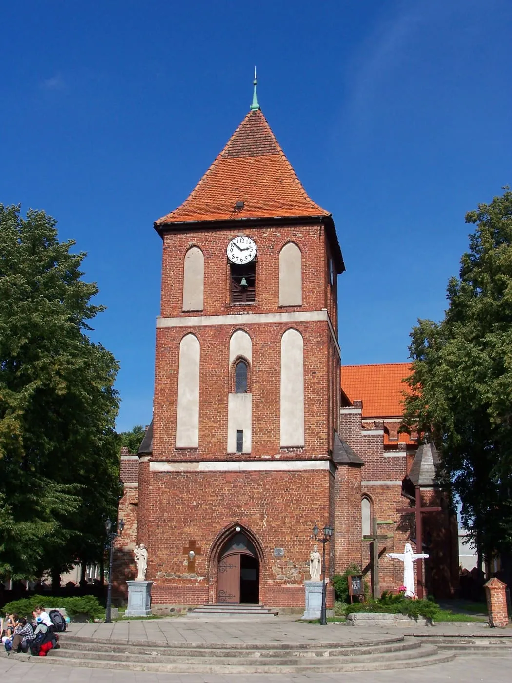 Ceglana, gotycka wieża z zegarem i czerwonym hełmem namiotowym, przed którą stoją figury na cokołach, na tle błękitnego nieba i drzew