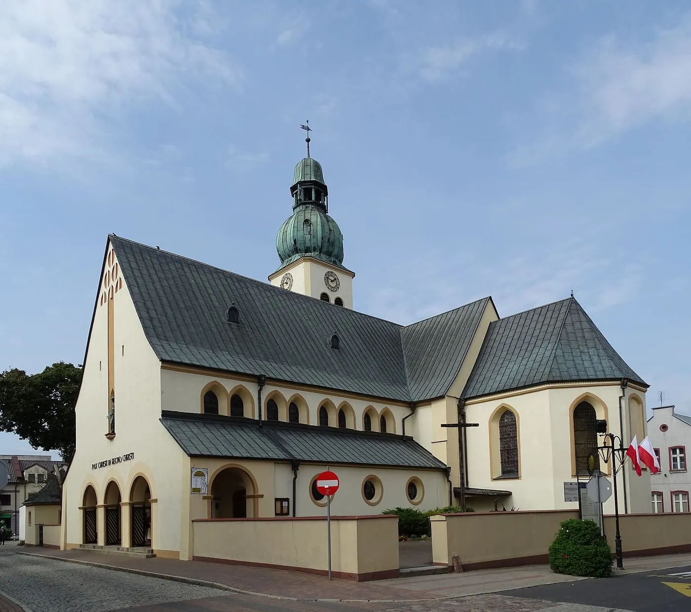 Jasno otynkowany kościół św. Jakuba w Człuchowie z szarymi dachami i wieżą z zielonym, cebulastym hełmem z latarnią, z arkadowym podcieniem i napisem „Pax Christi in regno Christi” nad wejściem