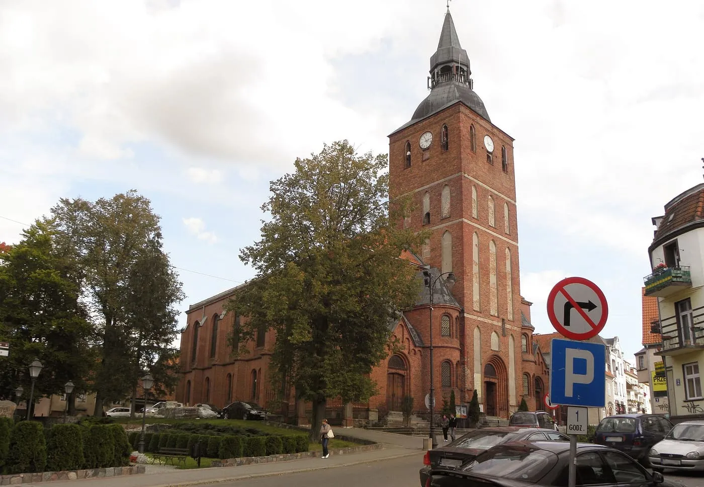 Kościół z czerwonej cegły z wysoką wieżą z zegarem i miedzianym hełmem, przy miejskiej ulicy z samochodami