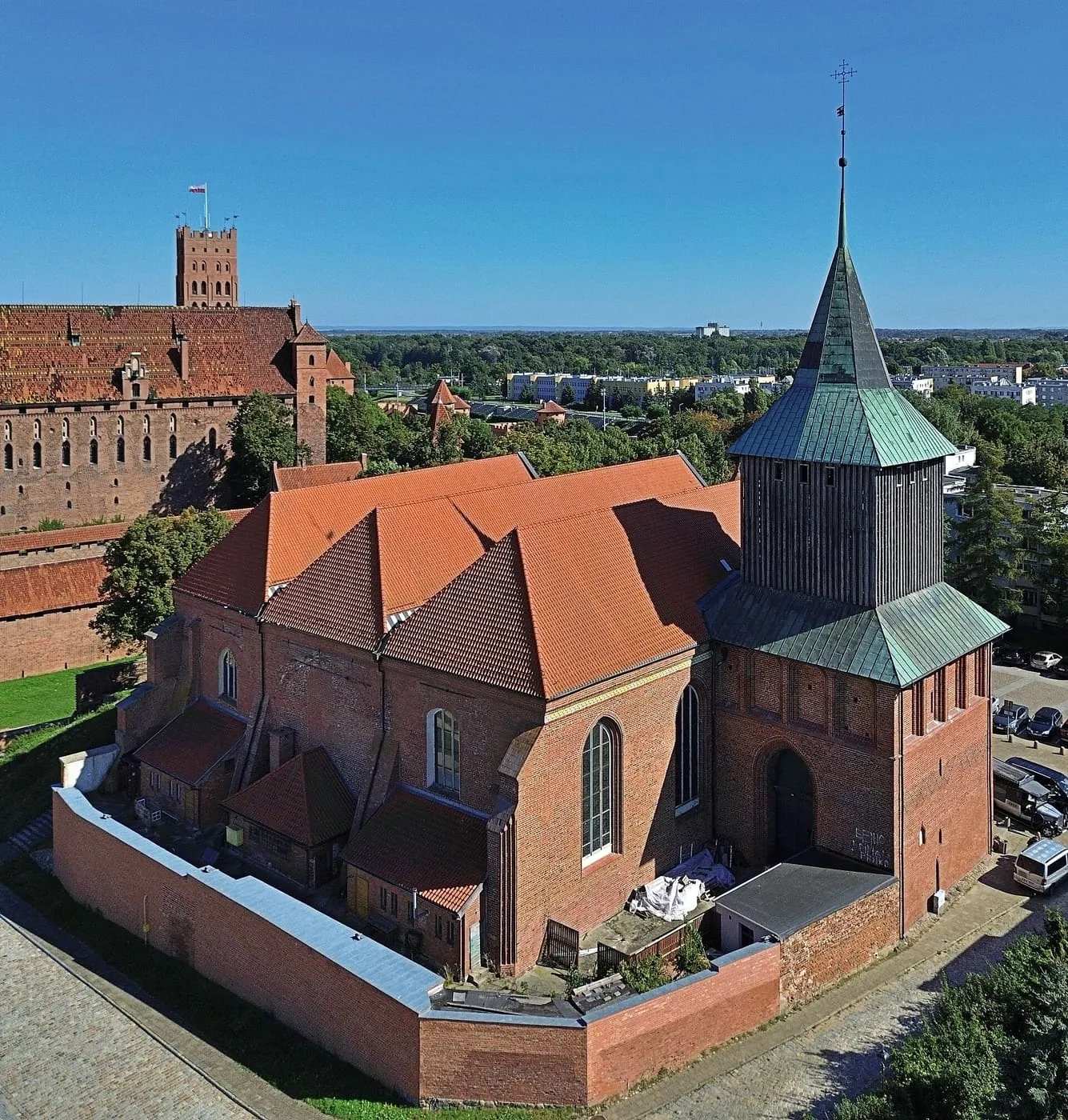 Widok z lotu ptaka na ceglany kościół św. Jana Chrzciciela w Malborku z osobno stojącą, drewnianą dzwonnicą, w tle mury zamku krzyżackiego