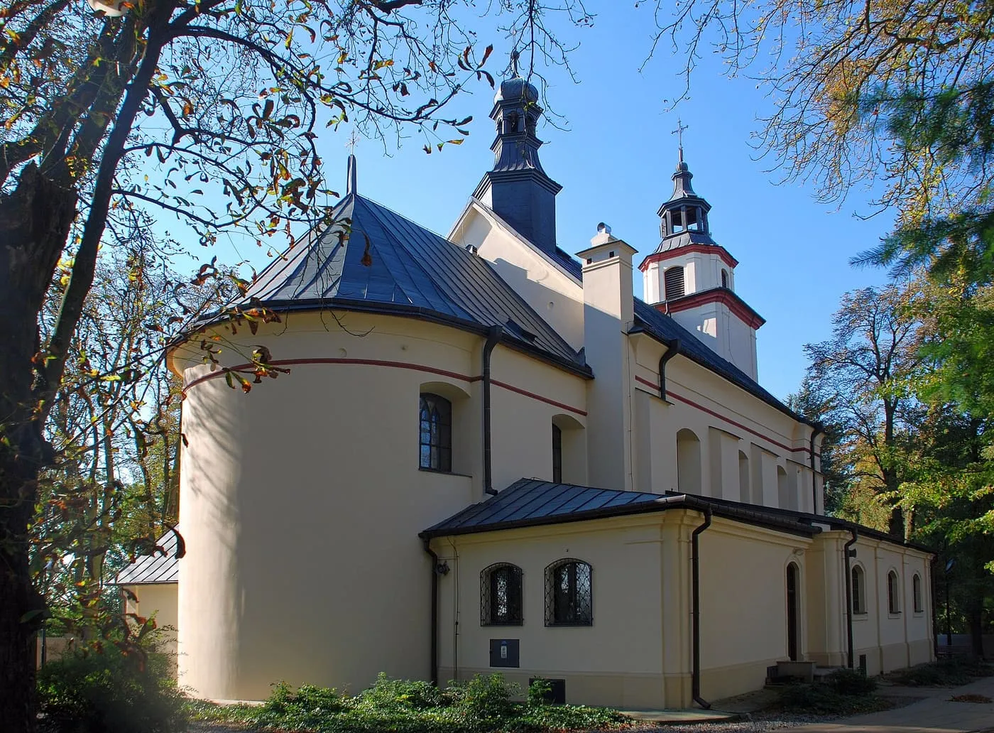 Kremowa, zaokrąglona apsyda kościoła z ciemnymi, blaszanymi dachami i wieżyczkami z baniastymi hełmami, widziana spod jesiennych drzew