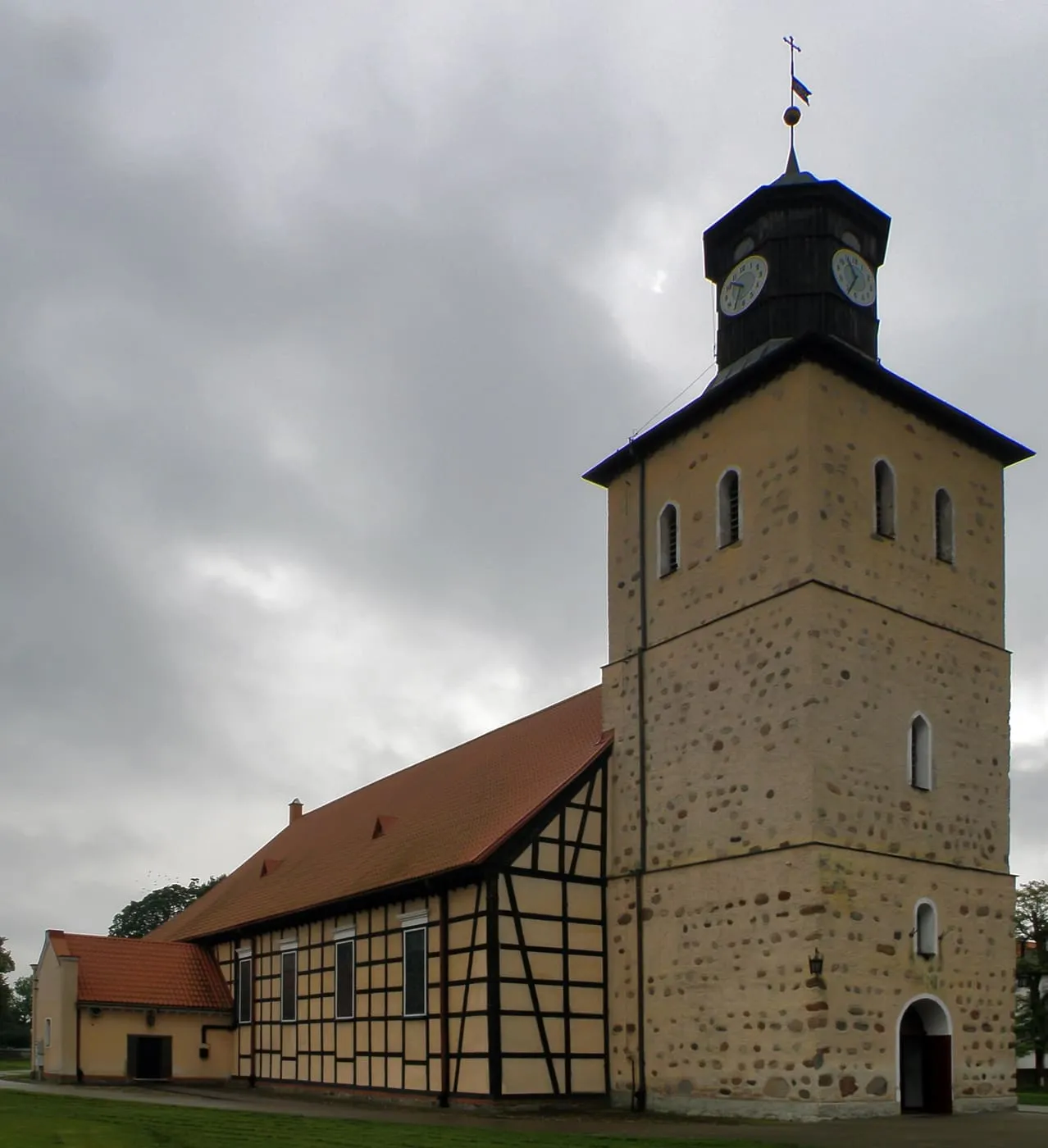 Ryglowa nawa kościoła w Piszu z czerwonym dachem, przylegająca do kamiennej, średniowiecznej wieży z drewnianą latarnią zegarową i małym hełmem zwieńczonym krzyżem, pod pochmurnym niebem