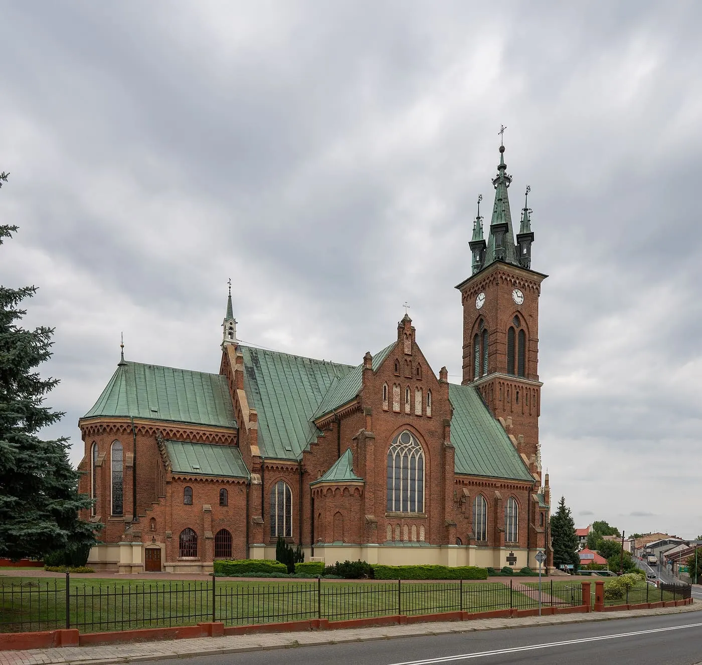 Neogotycki kościół z czerwonej cegły, z wysoką wieżą z zegarem i zielonymi, miedzianymi dachami, za czarnym ogrodzeniem przy ulicy, pod pochmurnym niebem