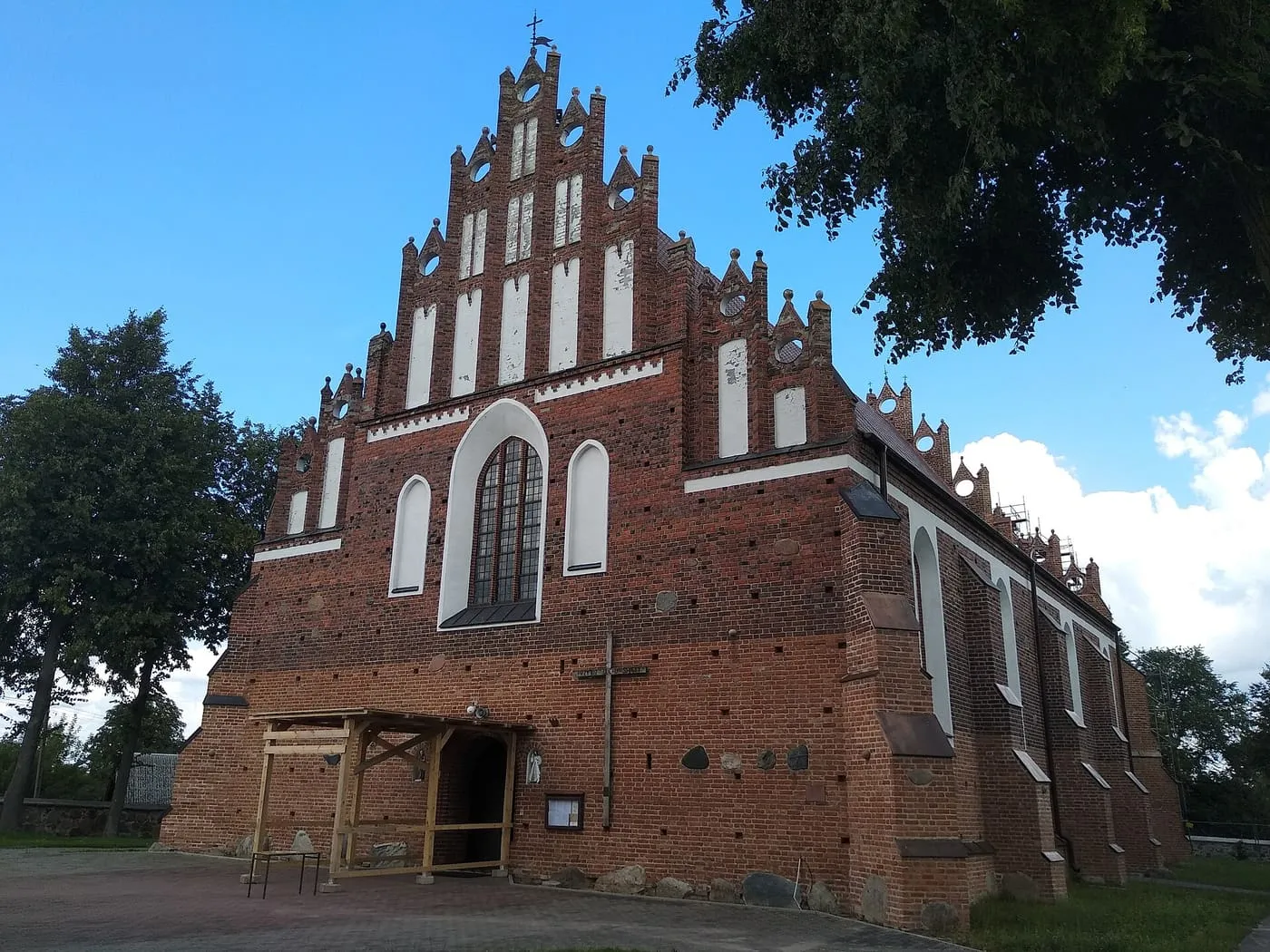 Ceglana, gotycka fasada zachodnia z wysokim, schodkowym szczytem i dużym ostrołukowym oknem, w cieniu drzew, pod błękitnym niebem, z drewnianym zadaszeniem przy wejściu