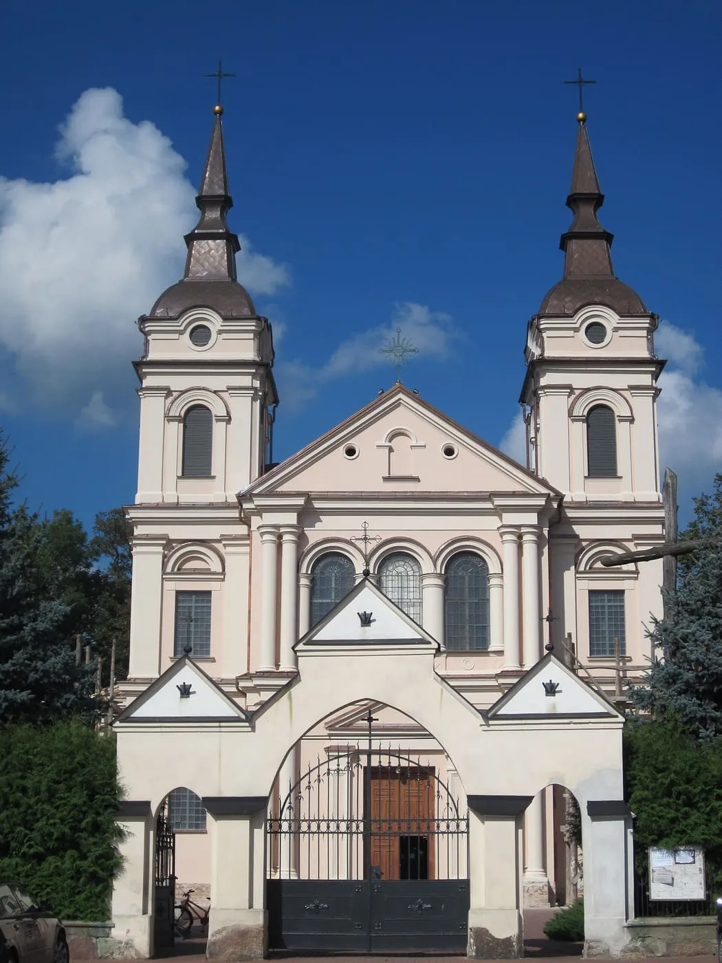 Dwuwieżowa, kremowa fasada kościoła z wieżami zwieńczonymi ciemnymi iglicami, a przed nią brama z żelaznymi wrotami, na tle błękitnego nieba