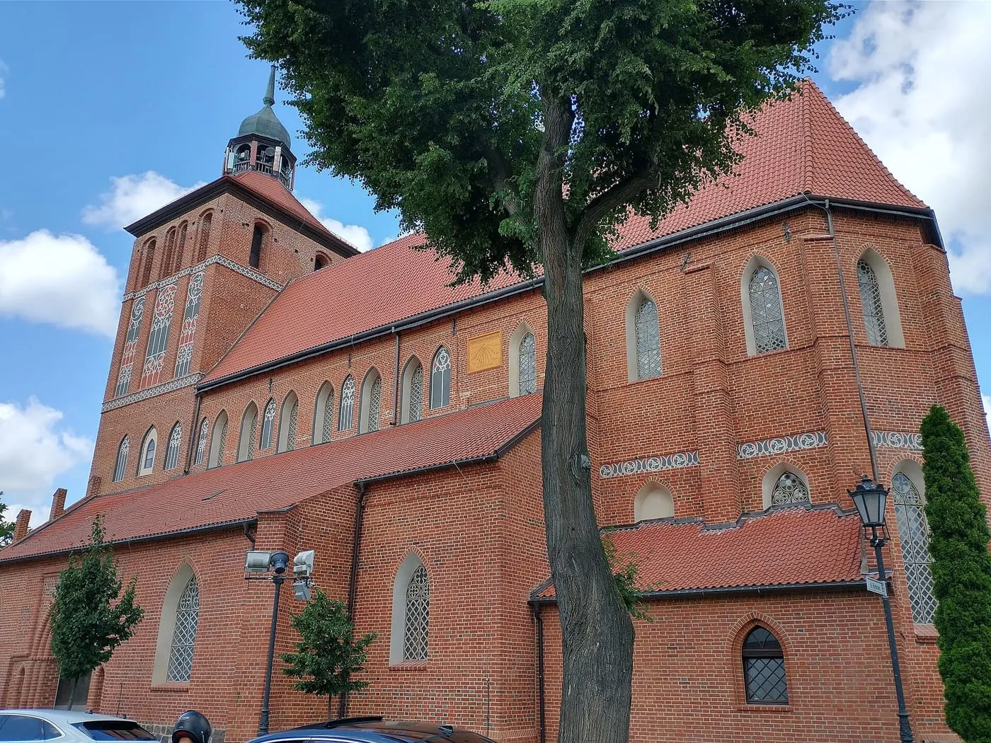 Ceglana, gotycka fara w Bartoszycach widziana z boku, z wysokim czerwonym dachem, oknami z maswerkami i wieżą ozdobioną białymi blendami, zwieńczoną miedzianym hełmem z latarnią, za drzewem przy ulicy