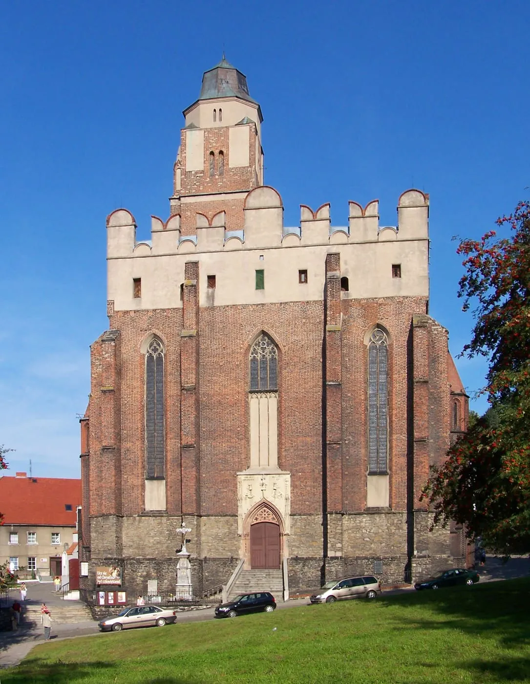 Masywna, ceglana fasada obronnego kościoła z gotyckimi oknami i attyką z blankami nad nawą, z wieżą wyrastającą z dachu; na pierwszym planie trawnik i samochody