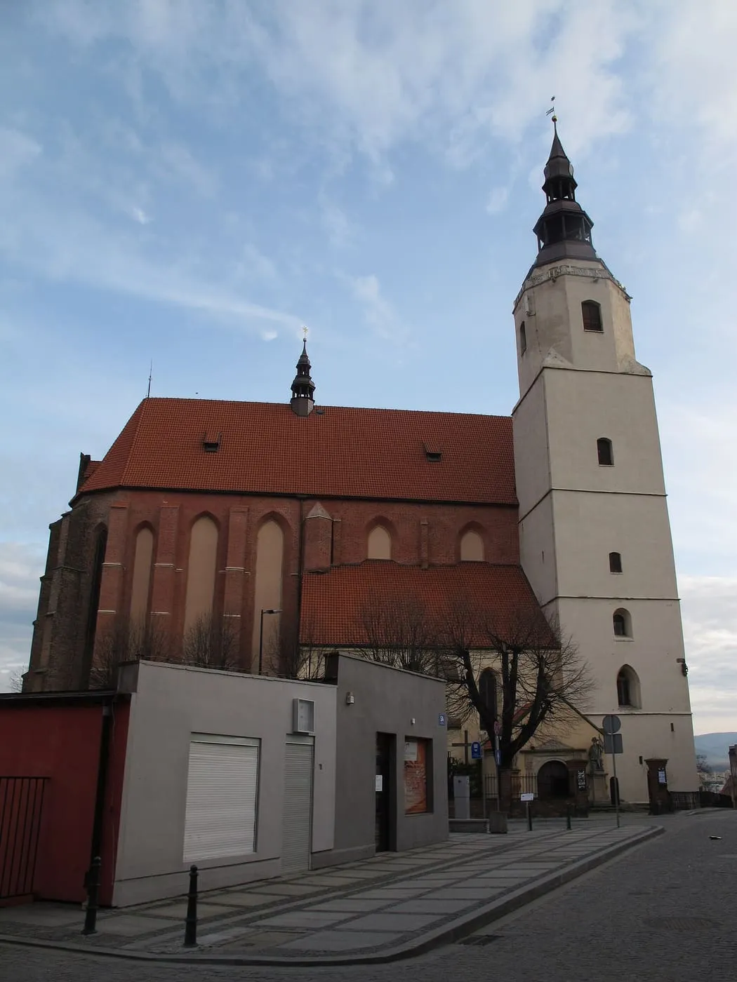 Gotycki kościół z czerwonej cegły z wysokim, czerwonym dachem i białą wieżą z ciemnym hełmem; na pierwszym planie brukowana ulica i szary, nowoczesny budynek