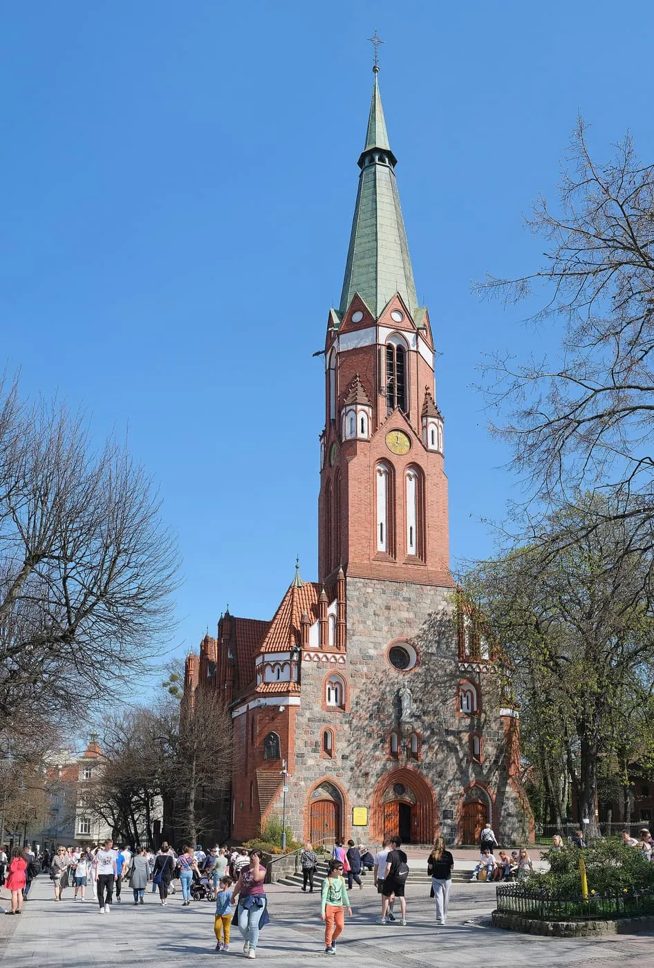 Neogotycki kościół z czerwonej cegły i kamienia z wysoką, smukłą wieżą zwieńczoną zieloną iglicą; przed wejściem spacerują ludzie, po bokach rosną bezlistne drzewa