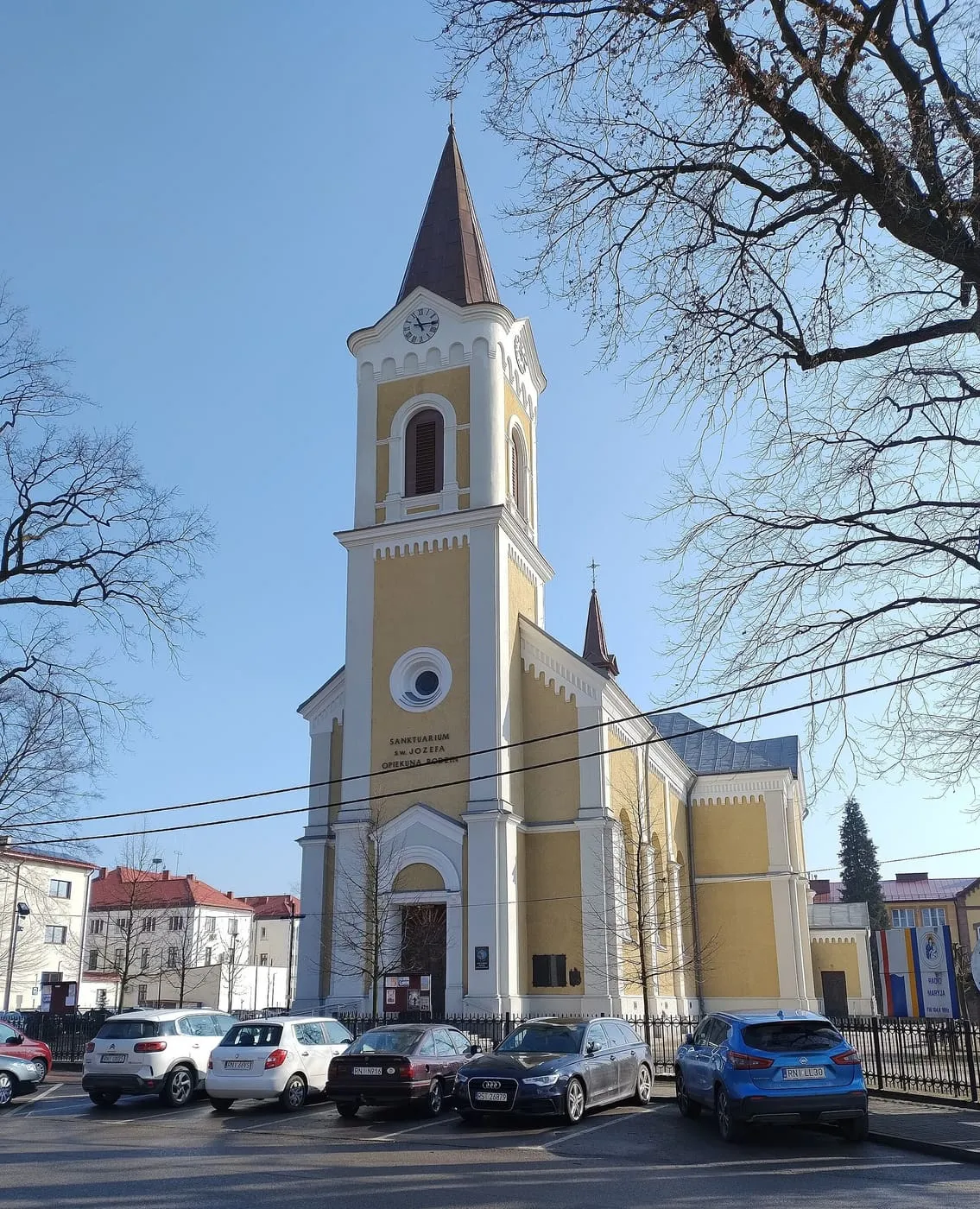 Żółto-kremowy kościół z wysoką wieżą z zegarem i ostrym hełmem, przed nim parking i drzewa bez liści