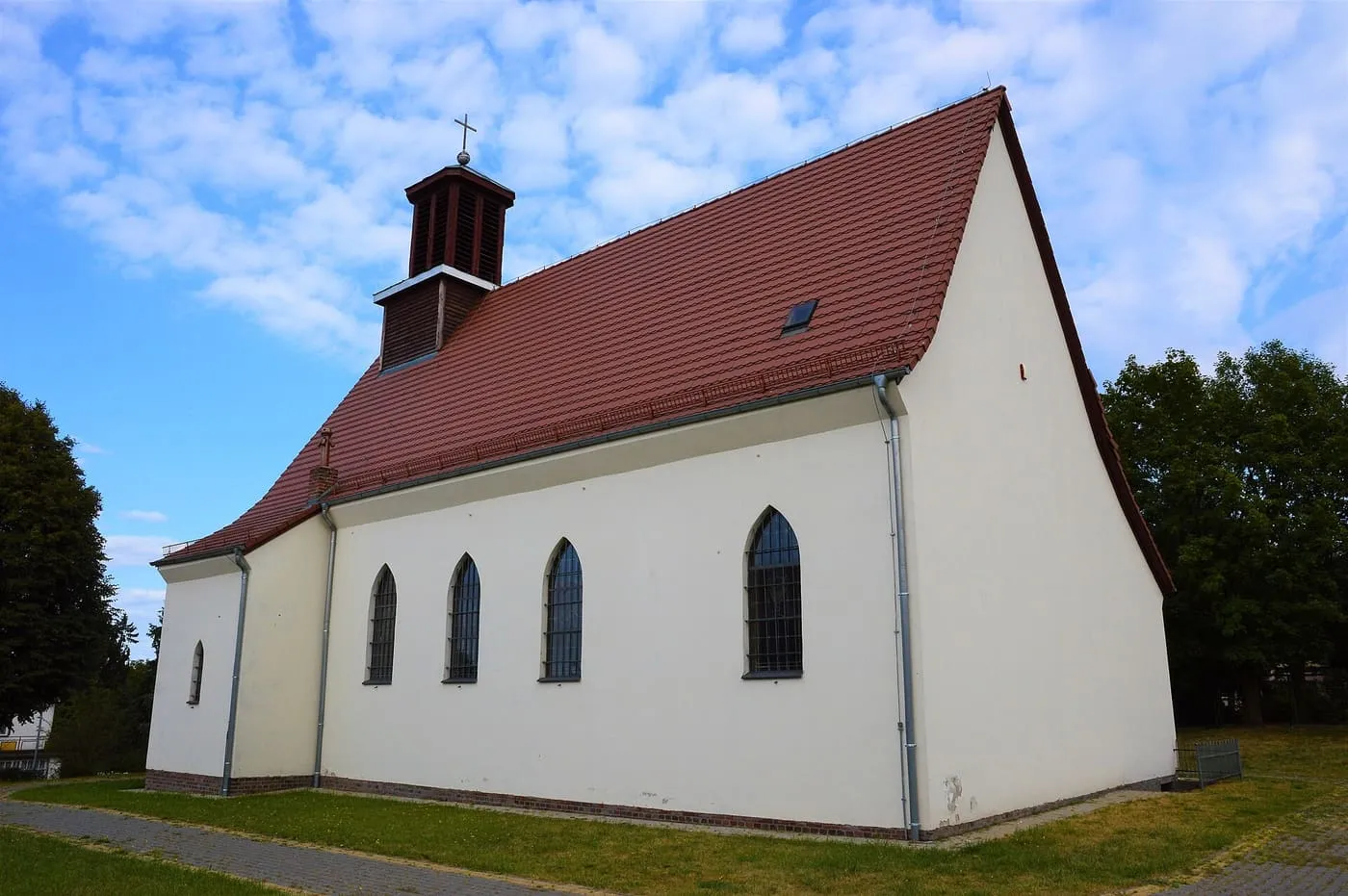 Biały kościół z czerwonym dachem, drewnianą wieżyczką na dachu i wąskimi, ostrołukowymi oknami, widziany z boku na tle chmur i drzew