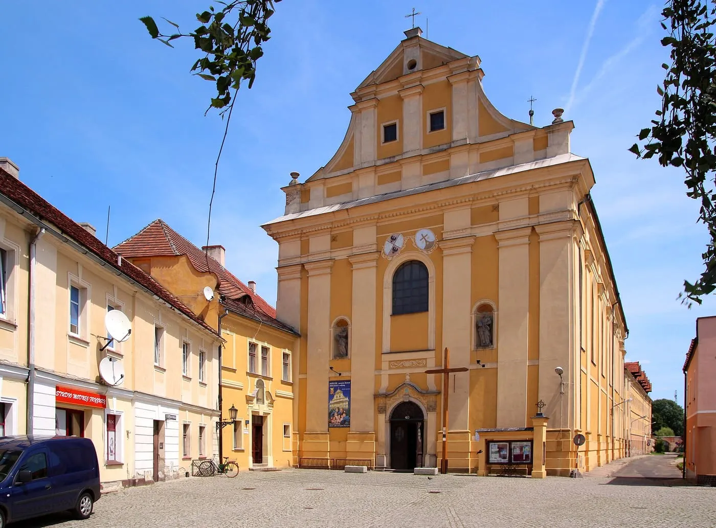 Żółta, barokowa fasada kościoła z wolutami i herbami, przed nią drewniany krzyż