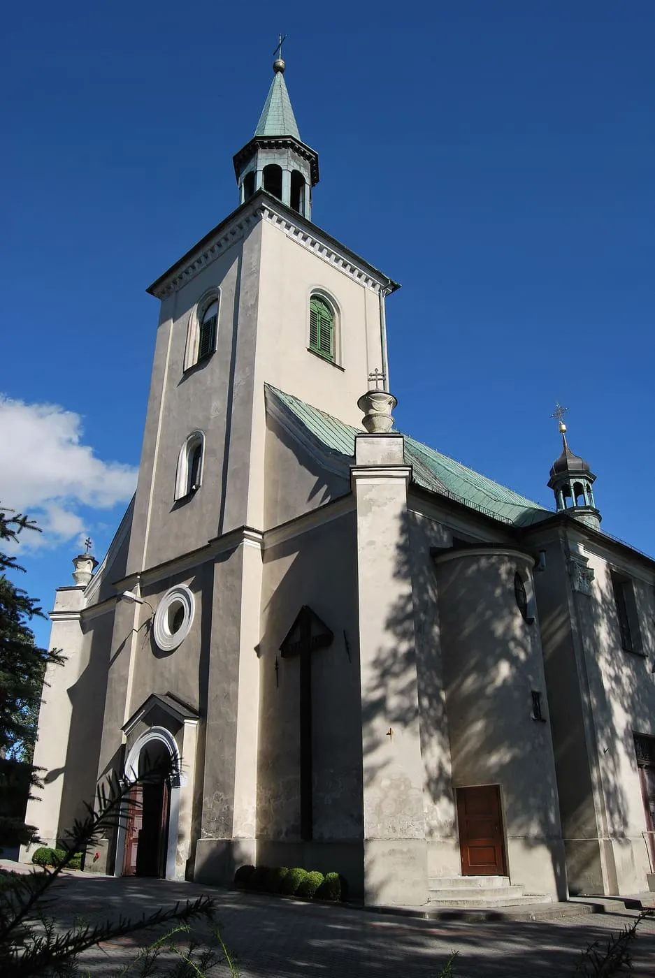Jasny, barokowy kościół z wysoką wieżą zwieńczoną zielonym hełmem z latarnią, widziany z ukosa od strony fasady na tle intensywnie błękitnego nieba