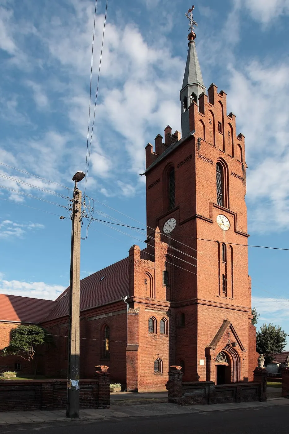 Ceglana wieża kościoła z zegarem, krenelowanym szczytem i iglicą, obok słup uliczny