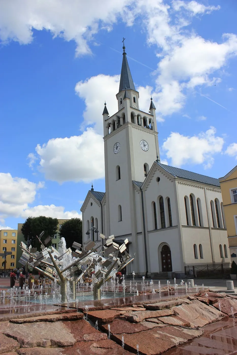 Jasna fasada kościoła św. Katarzyny w Bytowie z wysoką wieżą zegarową zwieńczoną szarą iglicą i czterema sterczynami, widziana z rynku z fontanną w pierwszym planie