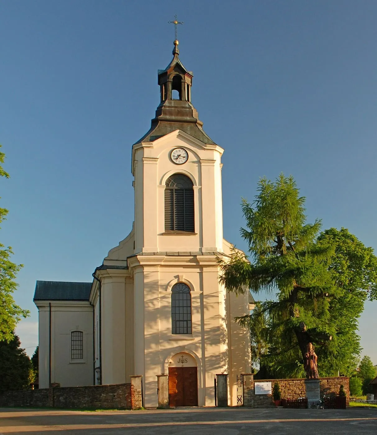 Jasnożółty kościół z wieżą z zegarem i baniastym hełmem, widziany od frontu w ciepłym świetle, obok wysokie drzewo