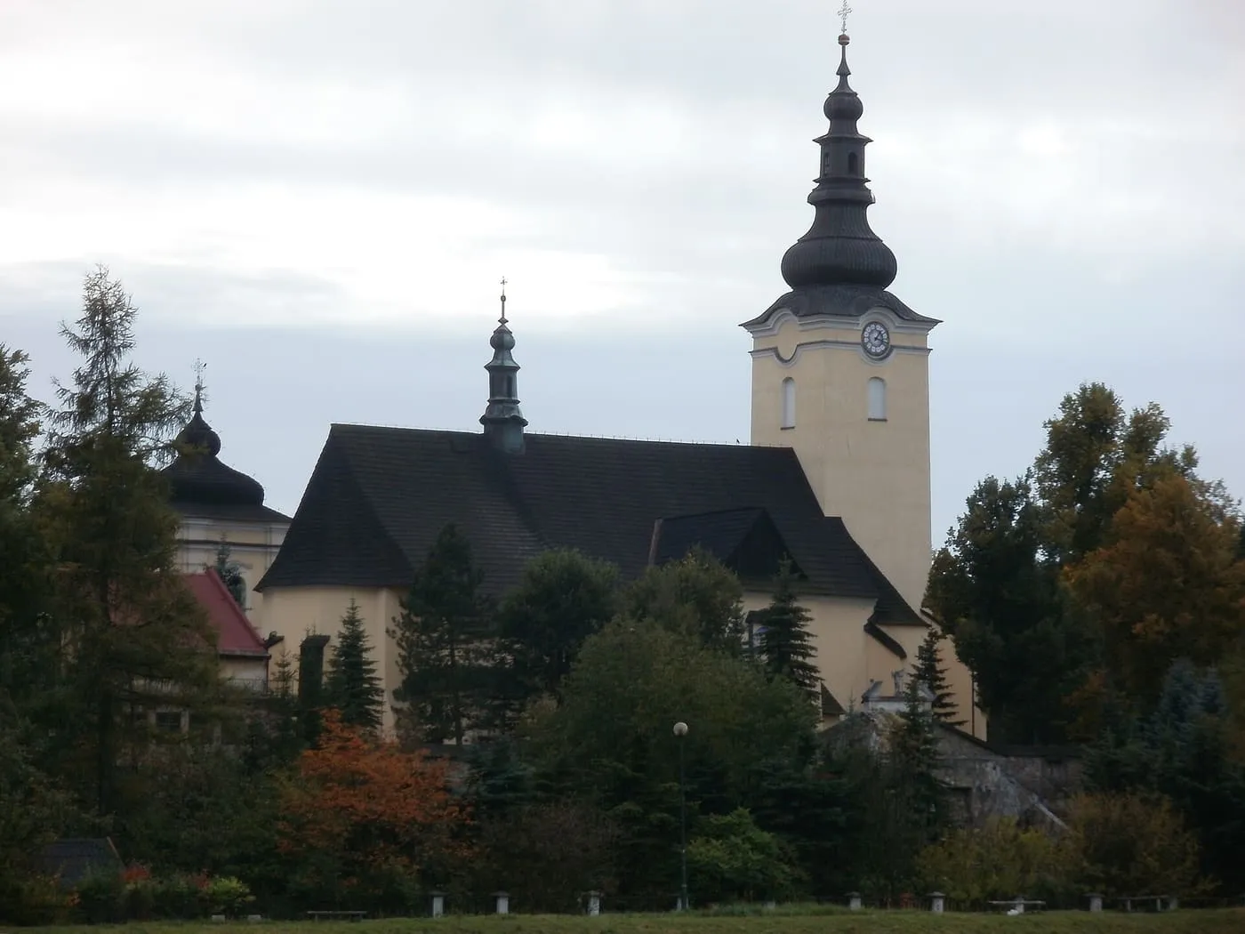 Kościół wśród jesiennych drzew, z ciemnym, stromym dachem nawy, jasną wieżą z zegarem zwieńczoną czarnym, baniastym hełmem i sygnaturką nad nawą