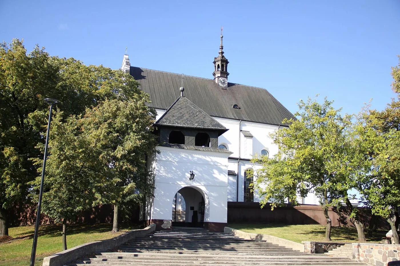 Biała fara z ciemnym dachem i sygnaturką z zegarem, a przed nią dzwonnica-brama z arkadowym przejściem i szerokie schody wśród zieleni drzew