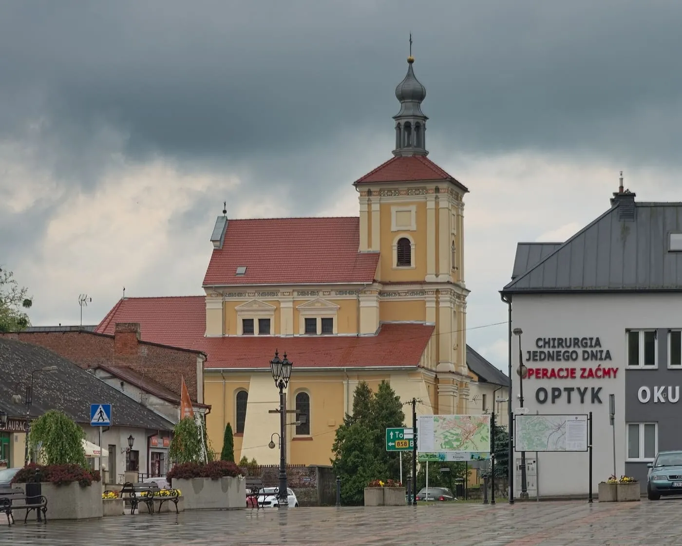 Żółty kościół z czerwonym dachem i wieżą zwieńczoną baniastym hełmem z latarnią, widziany z rynku; obok stoi nowoczesny budynek z szyldami, niebo jest zachmurzone