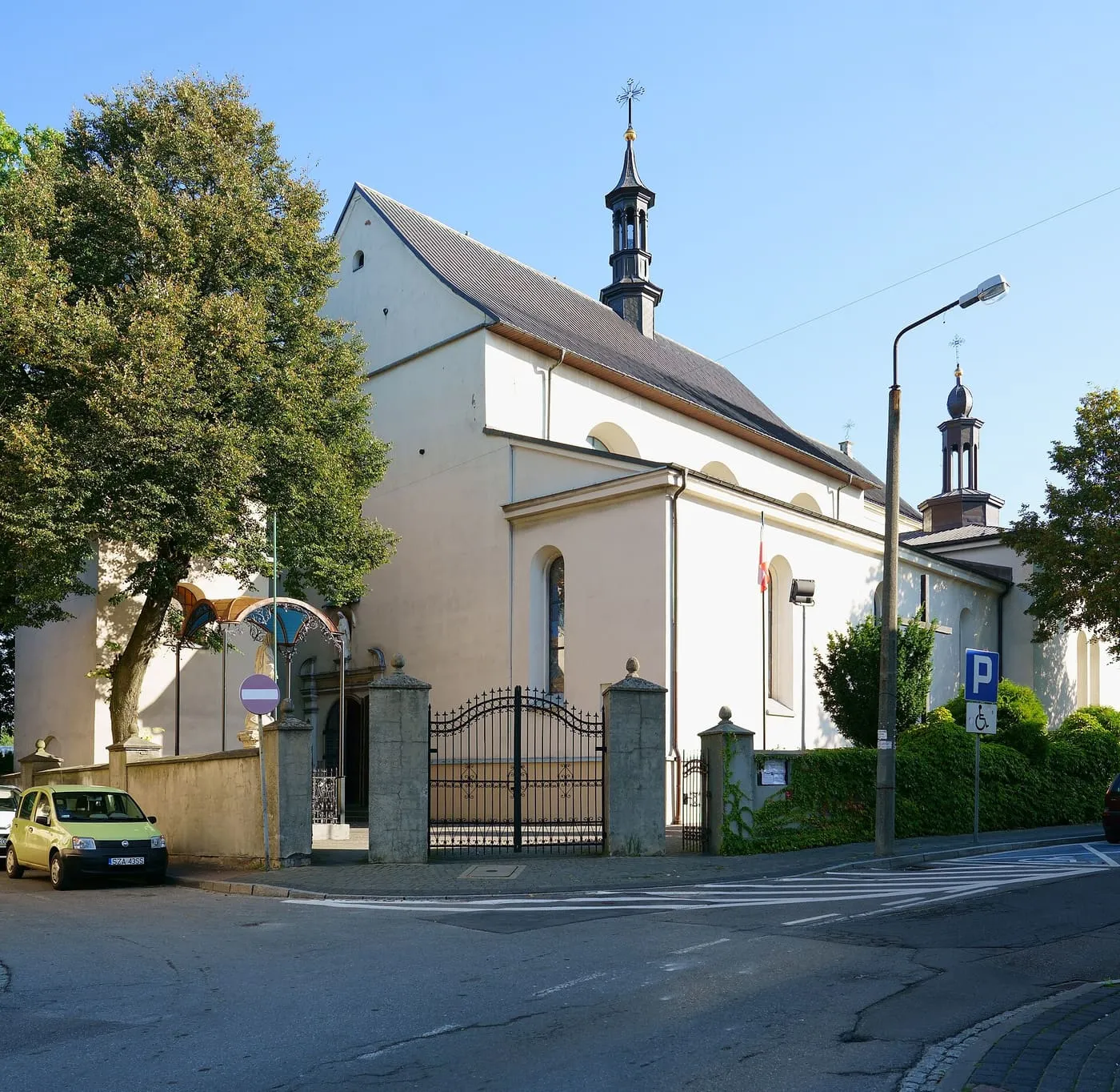 Biały, barokowy kościół z niewielką wieżyczką na sygnaturkę, widziany zza dużego drzewa i kutej bramy wjazdowej, z zaparkowanym samochodem przed wejściem