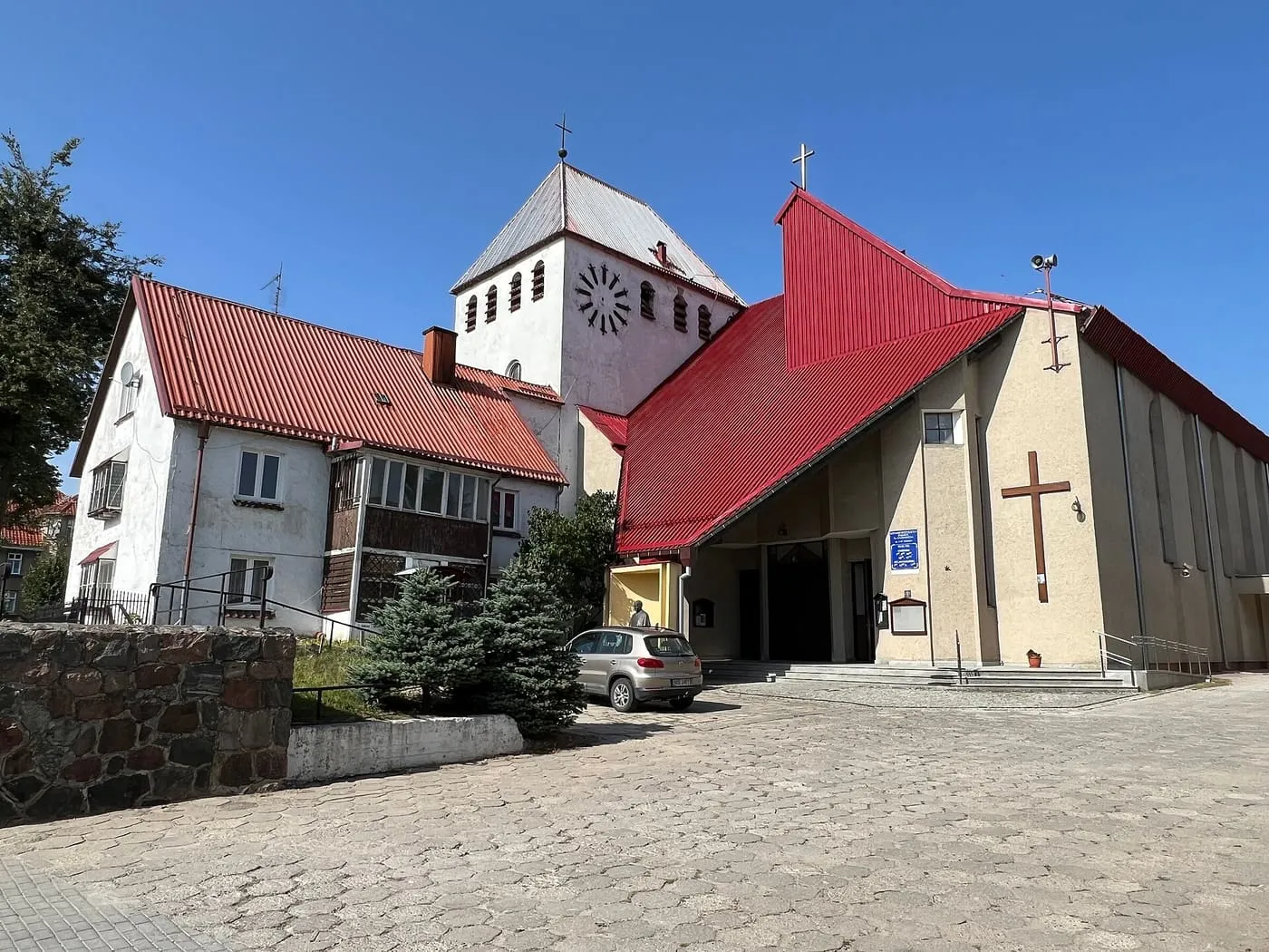 Współczesny kościół św. Kazimierza w Giżycku z czerwonym dachem nad wejściem i białą, kwadratową wieżą, w słoneczny dzień