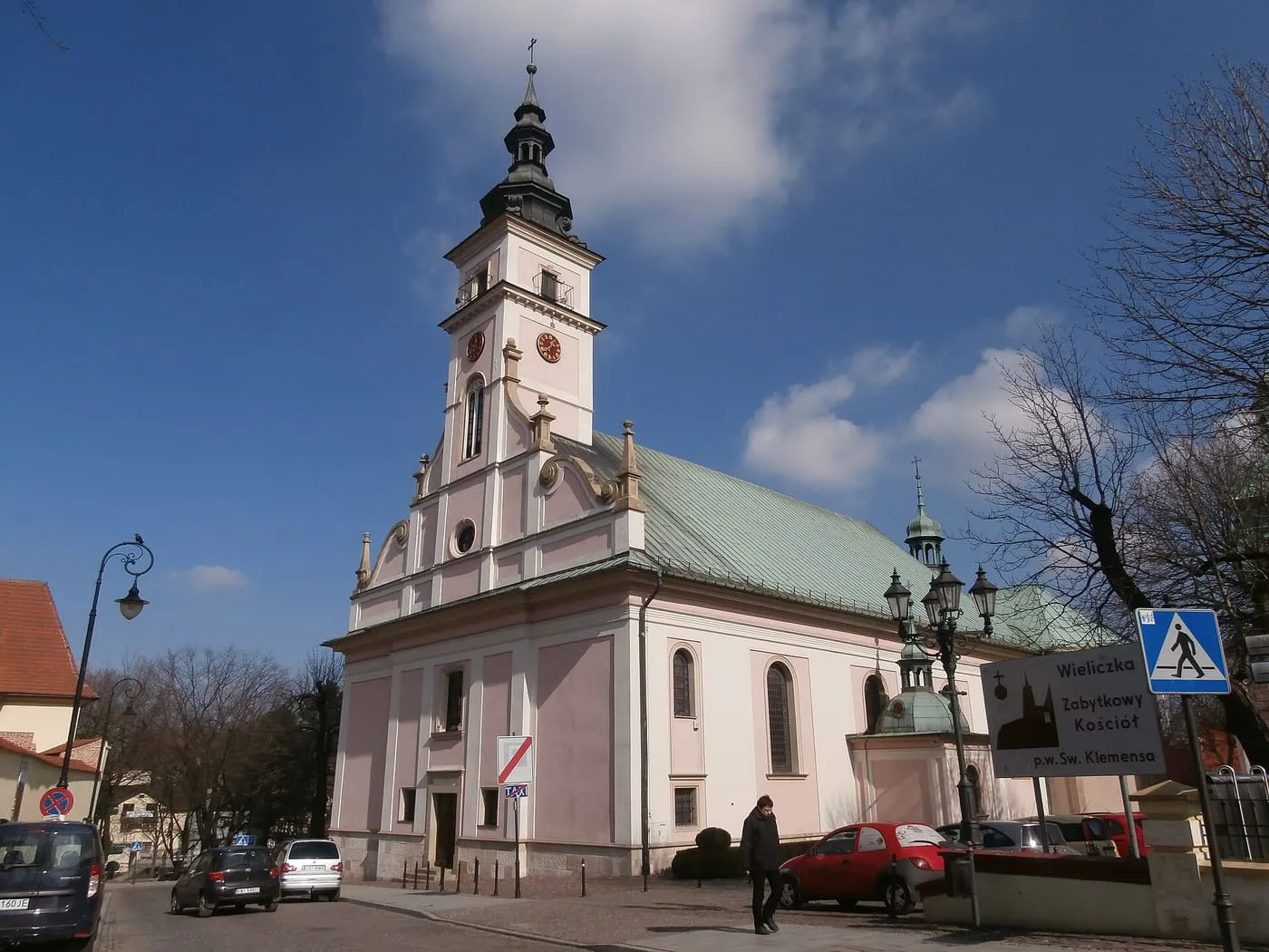 Różowo-biały, barokowy kościół z zielonym dachem i wieżą zegarową, przy ulicy tablica z nazwą zabytkowego kościoła