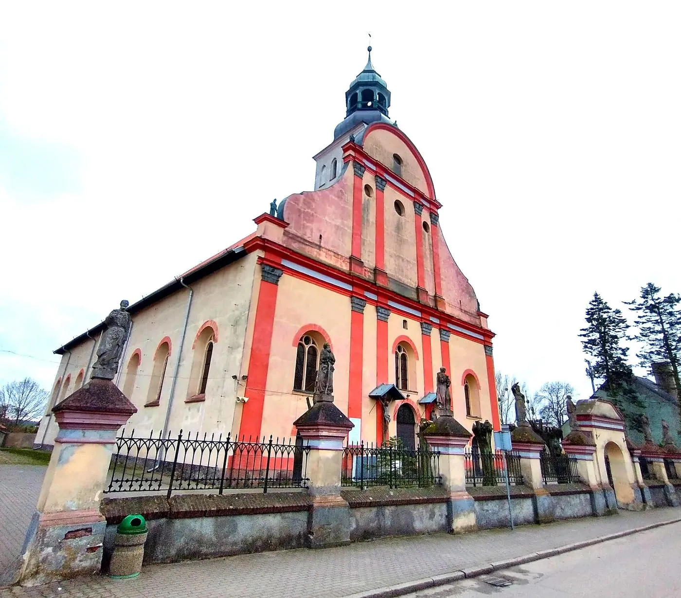 Późnobarokowy kościół z falistym szczytem fasady w czerwono-kremowych barwach i wieżyczką z hełmem, za ogrodzeniem z rzeźbami świętych na słupach
