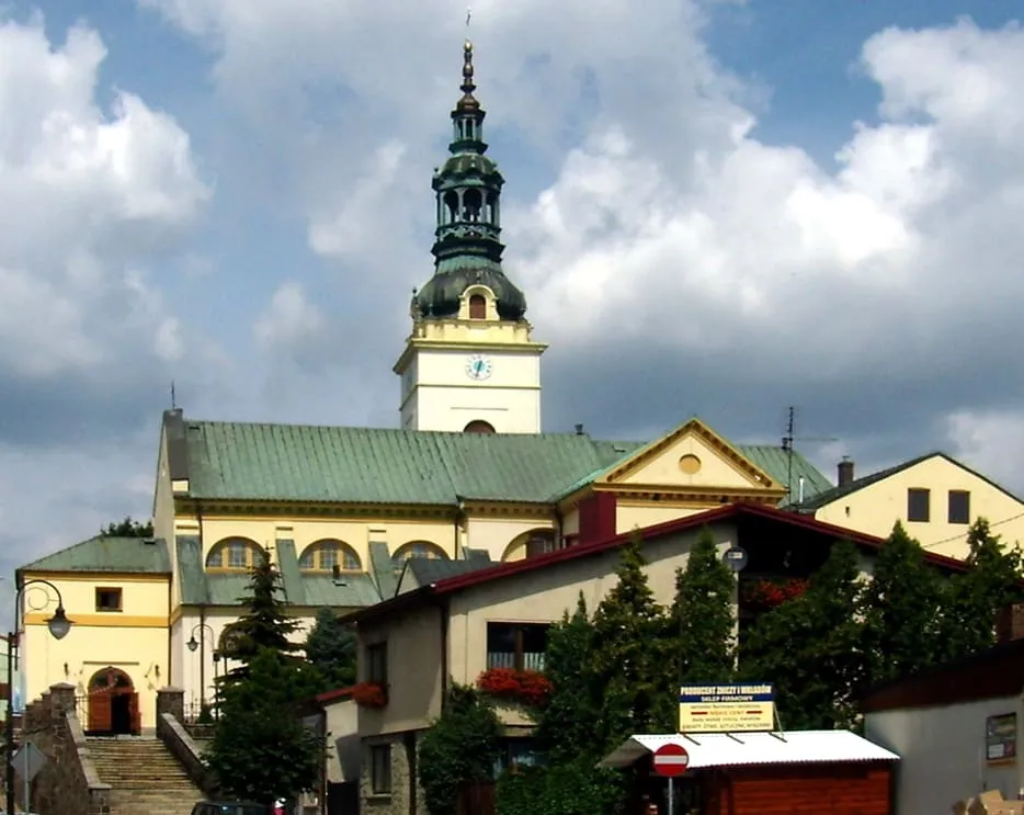 Jasnożółty kościół z zieloną wieżą zwieńczoną smukłym hełmem i zegarem, widziany od strony schodów prowadzących pod górę, otoczony niską zabudową miasteczka i drzewami, na tle zachmurzonego nieba