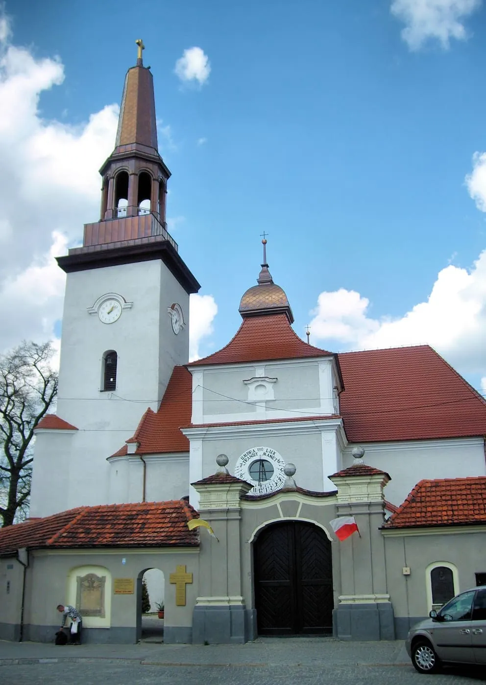 Biała wieża kościoła z zegarem i miedzianym hełmem, przed nią barokowa brama z czerwonym dachem i wielkimi ciemnymi wrotami; z prawej zaparkowany samochód