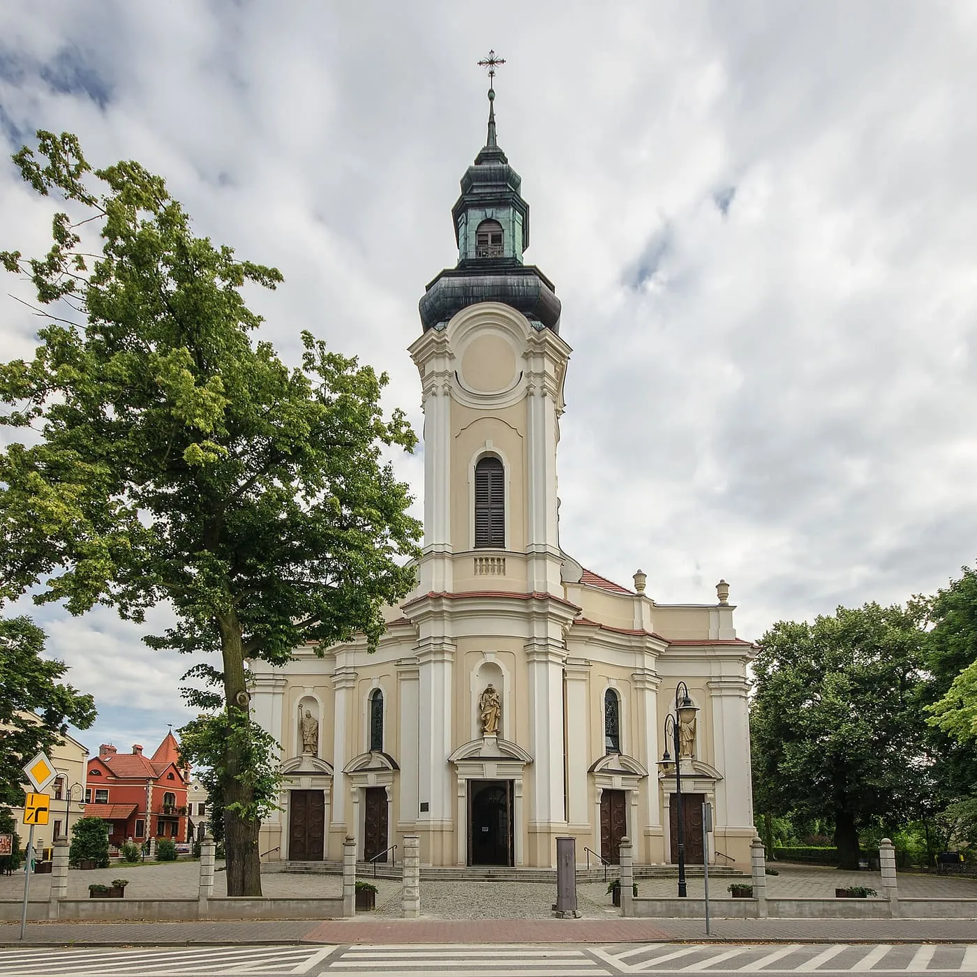 Neobarokowa fasada kościoła w kremowych barwach z wysoką wieżą zwieńczoną ciemnym hełmem, stojąca przy placu; z lewej rozłożyste drzewo