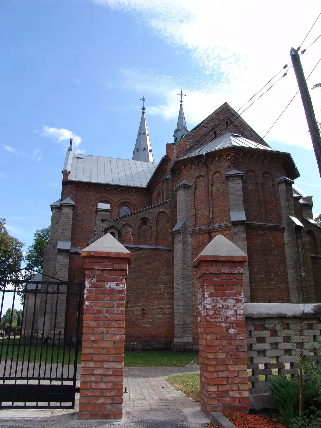 Ceglane ściany i wieżyczki kościoła widoczne zza ceglanej bramy z kamiennymi zwieńczeniami słupów, na tle błękitnego nieba