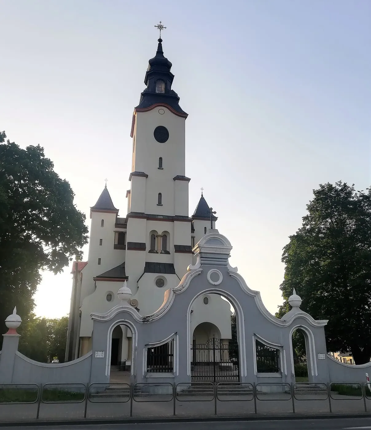 Biały kościół z wieżą z zegarem i ciemnym hełmem, przed nim ozdobna, faliście zakończona brama, w tle drzewa i niebo