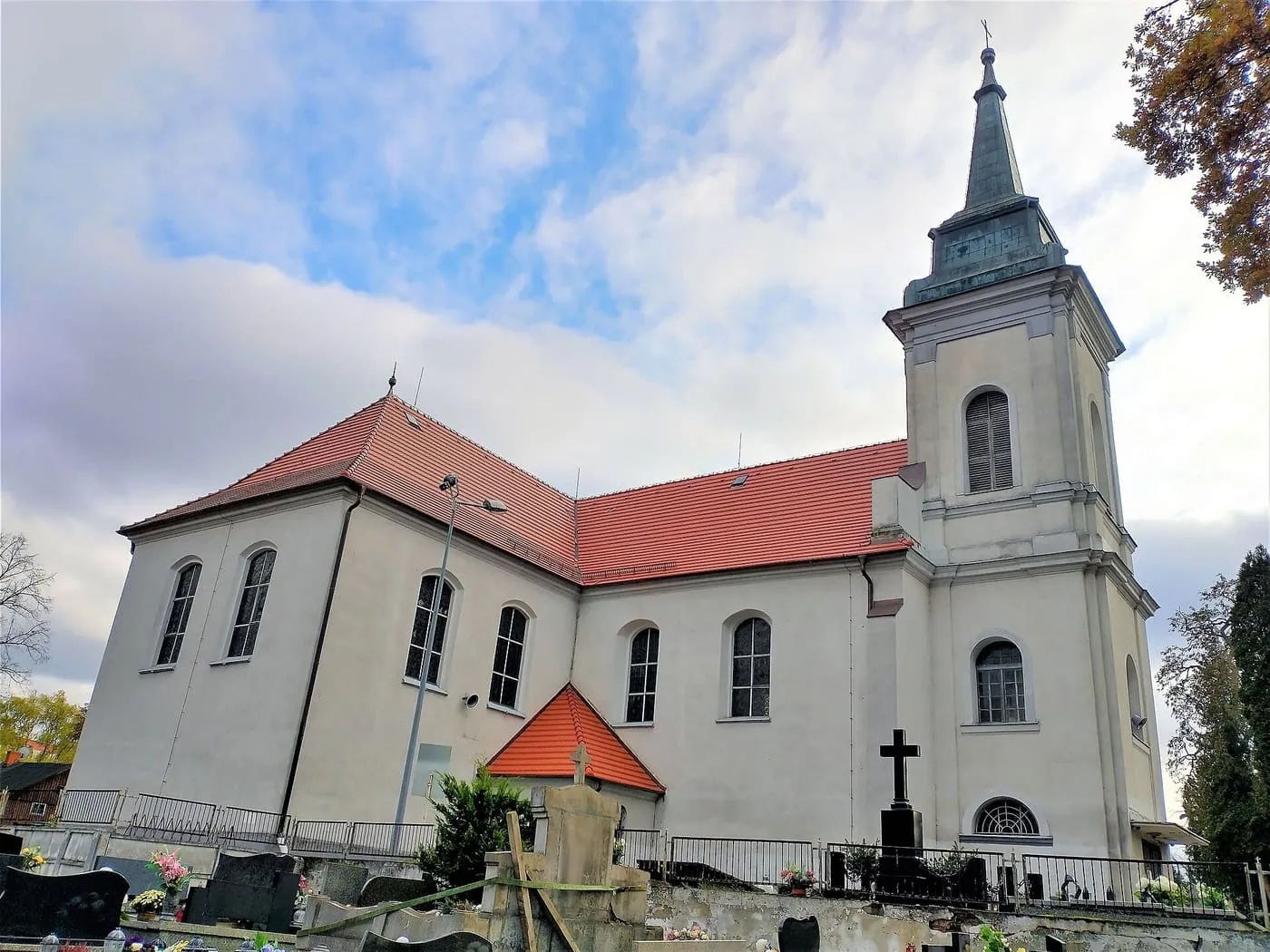 Jasny kościół z czerwonym dachem i wysoką wieżą zwieńczoną smukłym, miedzianym hełmem, otoczony przykościelnym cmentarzem z krzyżami, pod zachmurzonym niebem