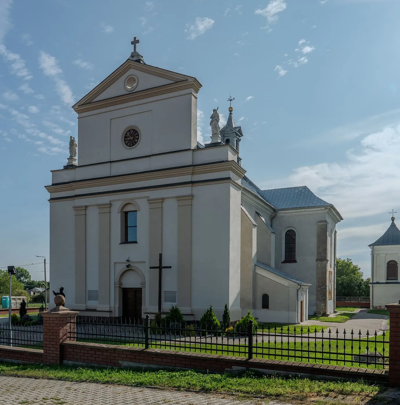 Jasna fasada barokowego kościoła z trójkątnym frontonem i okrągłym okienkiem, z rzeźbami na narożnikach; przed nim ogrodzenie z ceglanymi słupkami i trawnik
